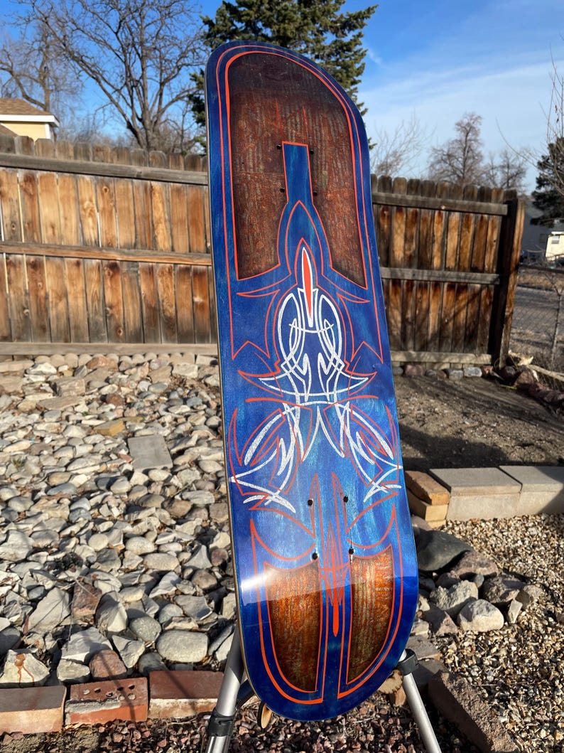 May include: A skateboard deck featuring a dark wood grain finish with a blue metallic overlay. The surface is decorated with intricate orange and white pinstripe designs in a symmetrical pattern, including thin lines and pointed shapes.