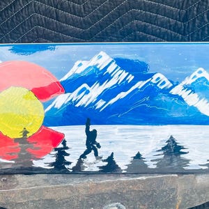May include: A skateboard deck featuring a painted landscape of snow-capped blue mountains, a dark blue sky, and a foreground of black pine trees. A large red and yellow Colorado state flag emblem is on the left, with a black silhouette of Bigfoot.