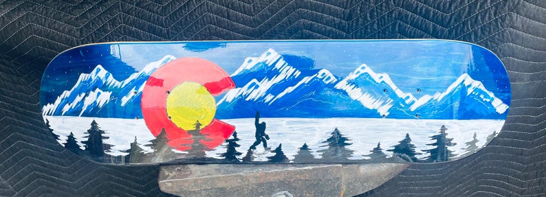 May include: A skateboard deck featuring a painted landscape of snow-capped blue mountains, a dark blue sky, and a foreground of black pine trees. A large red and yellow Colorado state flag emblem is on the left, with a black silhouette of Bigfoot.