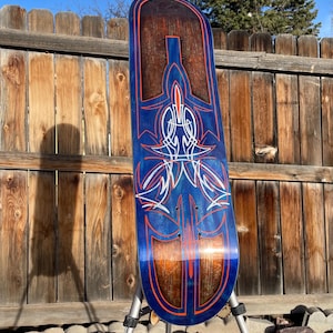 May include: A skateboard deck featuring a deep blue finish with a visible wood grain texture. The surface is decorated with intricate pinstriping in bright orange and white, forming symmetrical, flowing patterns that run along the length.