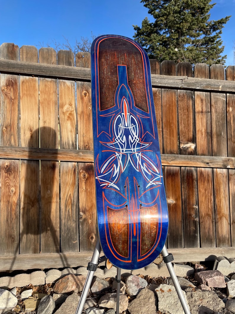 May include: A skateboard deck featuring a deep blue finish with a visible wood grain texture. The surface is decorated with intricate pinstriping in bright orange and white, forming symmetrical, flowing patterns that run along the length.