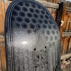 May include: A black skateboard deck featuring a prominent silver glitter finish and a dark grey honeycomb pattern printed across the upper section. The deck includes pre-drilled mounting holes for trucks and a distinct outlined graphic design.