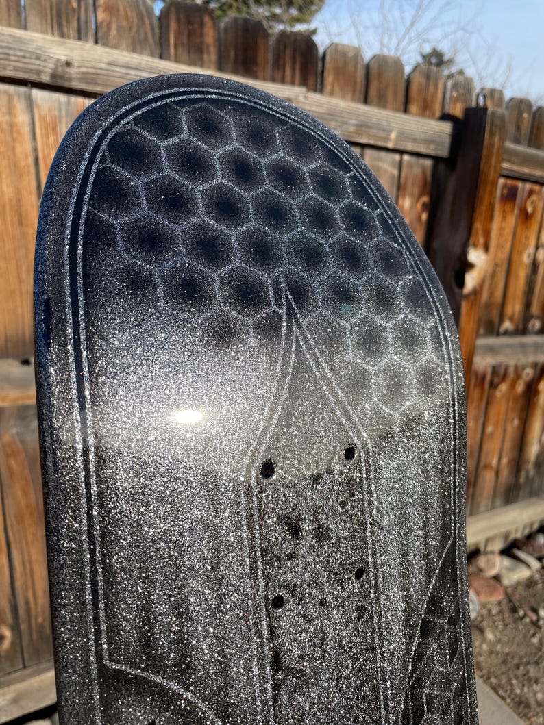 May include: A black skateboard deck featuring a prominent silver glitter finish and a dark grey honeycomb pattern printed across the upper section. The deck includes pre-drilled mounting holes for trucks and a distinct outlined graphic design.