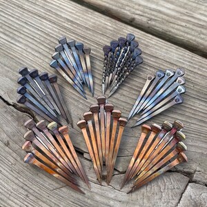 Op de afbeelding: Een assortiment metalen hoefijzernagels gerangschikt in waaiervormige patronen. De nagels vertonen een reeks kleuren, waaronder brons, blauw en zilver, met getextureerde koppen. De opstelling staat op een verweerd houten oppervlak.