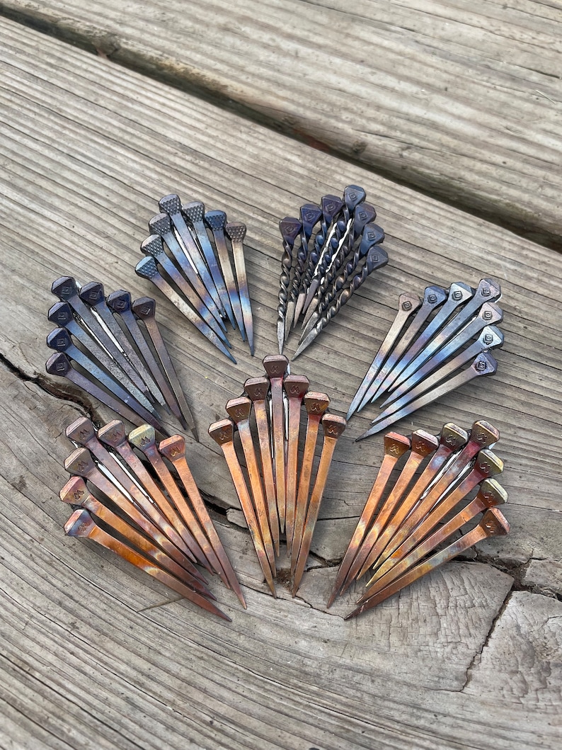 Op de afbeelding: Een assortiment metalen hoefijzernagels gerangschikt in waaiervormige patronen. De nagels vertonen een reeks kleuren, waaronder brons, blauw en zilver, met getextureerde koppen. De opstelling staat op een verweerd houten oppervlak.