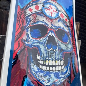 May include: A skateboard deck featuring a vibrant graphic illustration of a skull wearing a feathered headdress. The artwork uses bold red, blue, and white colours with detailed line work, depicting the skull with dark eye sockets and teeth.