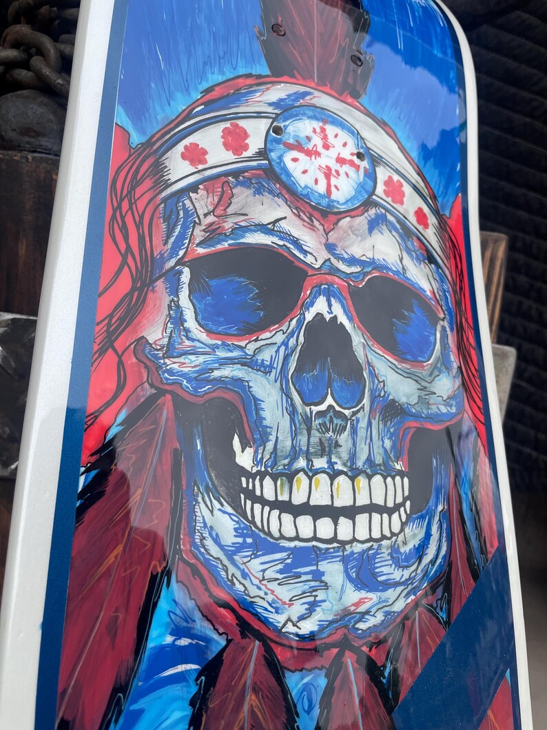 May include: A skateboard deck featuring a vibrant graphic illustration of a skull wearing a feathered headdress. The artwork uses bold red, blue, and white colours with detailed line work, depicting the skull with dark eye sockets and teeth.