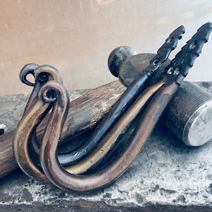 May include: Two hand-forged metal hooks with a curved, scroll-like design at the top and a textured, tapered end. The hooks feature a dark, burnished bronze and black finish, showcasing a rustic, hammered appearance with visible metalwork details.