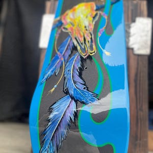 May include: A skateboard deck featuring a black centre panel with a painted illustration of a multi-coloured animal skull and blue and purple feathers. The deck is framed by bright blue edges with green pinstriping and a glossy finish.