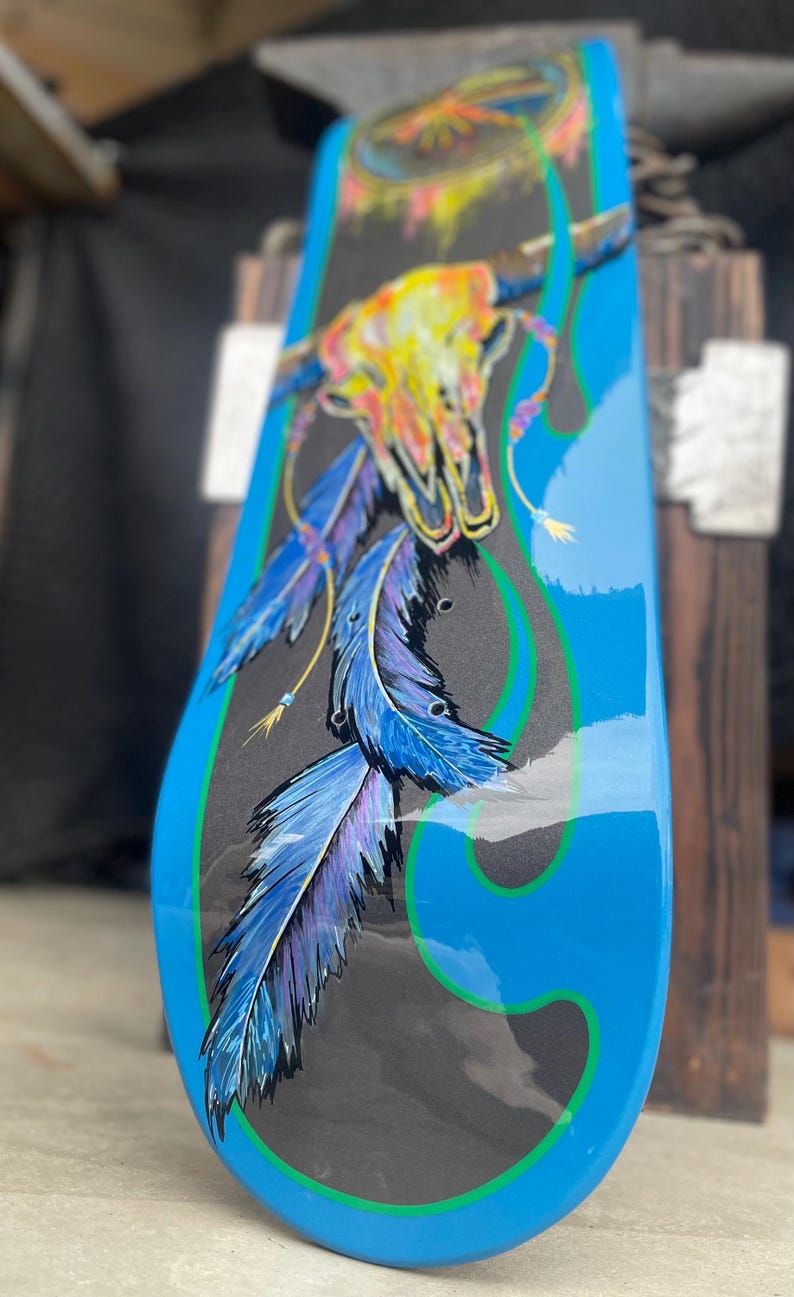 May include: A skateboard deck featuring a black centre panel with a painted illustration of a multi-coloured animal skull and blue and purple feathers. The deck is framed by bright blue edges with green pinstriping and a glossy finish.