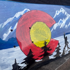 May include: A skateboard deck featuring a hand-painted scene of snow-capped blue mountains and a forest. A large red letter C with a yellow centre circle is prominently displayed in the foreground, with a silhouette of a creature walking among trees.