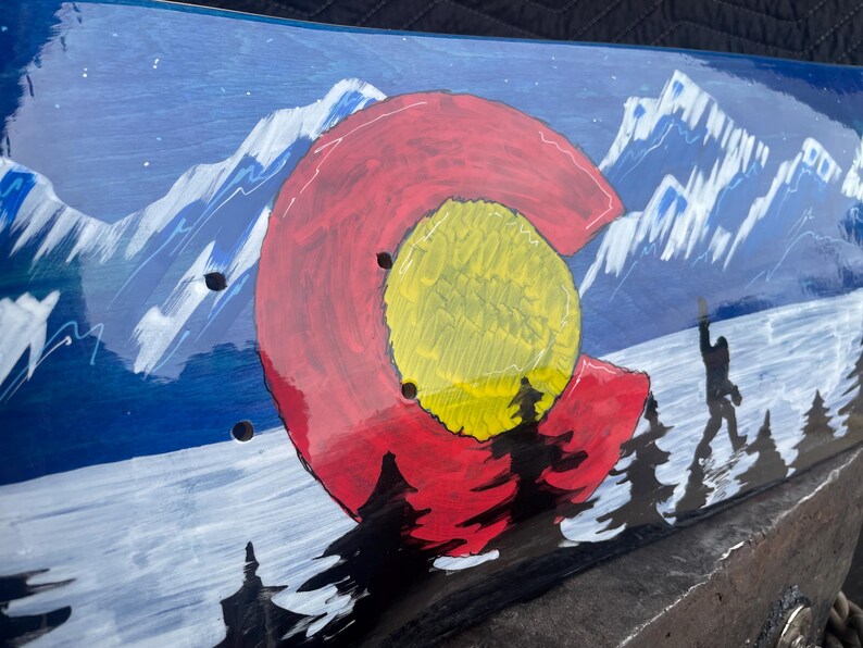 May include: A skateboard deck featuring a hand-painted scene of snow-capped blue mountains and a forest. A large red letter C with a yellow centre circle is prominently displayed in the foreground, with a silhouette of a creature walking among trees.