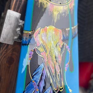 May include: A skateboard deck featuring a painted steer skull in yellow and pink tones, accented with blue and purple feathers and beaded strands. The background is a dark grey with bright blue side borders and a circular dreamcatcher design.