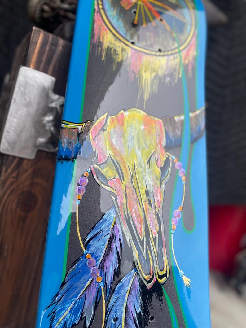 May include: A skateboard deck featuring a painted steer skull in yellow and pink tones, accented with blue and purple feathers and beaded strands. The background is a dark grey with bright blue side borders and a circular dreamcatcher design.