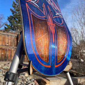 May include: A skateboard deck featuring a deep blue metallic finish with intricate orange pinstriping and white line art. The centre displays two large, symmetrical panels with a textured, amber-toned wood grain effect under a glossy clear coat.