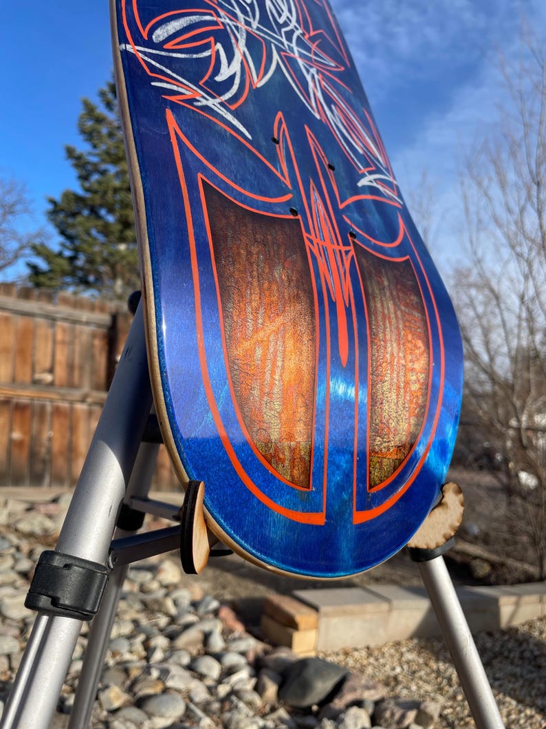 May include: A skateboard deck featuring a deep blue metallic finish with intricate orange pinstriping and white line art. The centre displays two large, symmetrical panels with a textured, amber-toned wood grain effect under a glossy clear coat.