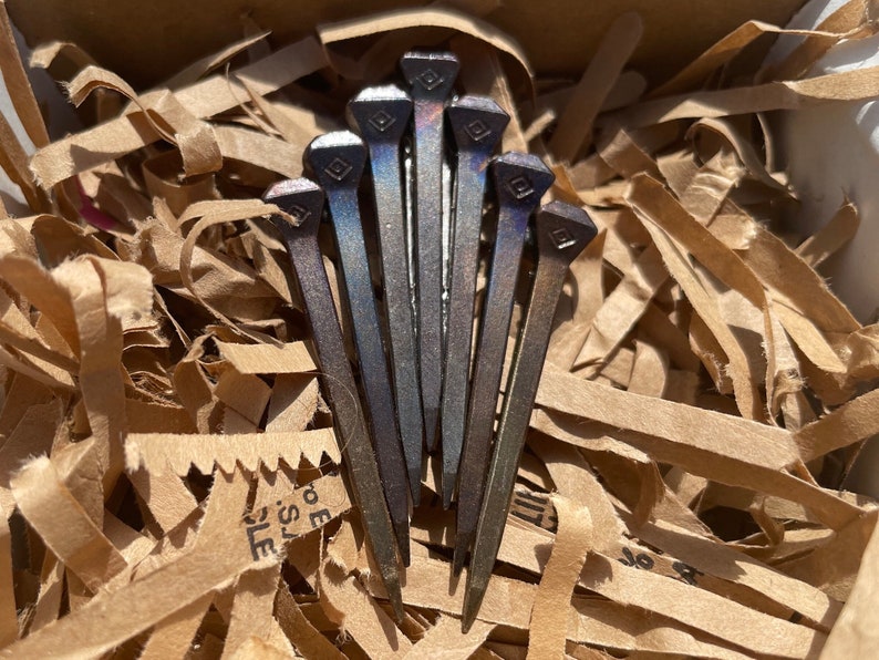 Op de afbeelding: Een verzameling van zeven donkere metalen hoefijzernagels met vierkante koppen, gerangschikt in een waaier. De nagels hebben een licht iriserende afwerking en zijn genesteld in versnipperd bruin papier in een kartonnen doos.