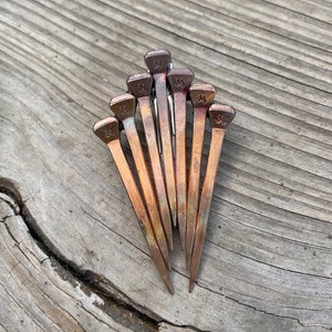 Op de afbeelding: Een verzameling van negen koperkleurige hoefijzernagels, gerangschikt in een waaier. Elke nagel heeft een rechthoekige kop met een decoratief ontwerp. De nagels liggen op een verweerd, grijs houten oppervlak met zichtbare nerven en scheuren.