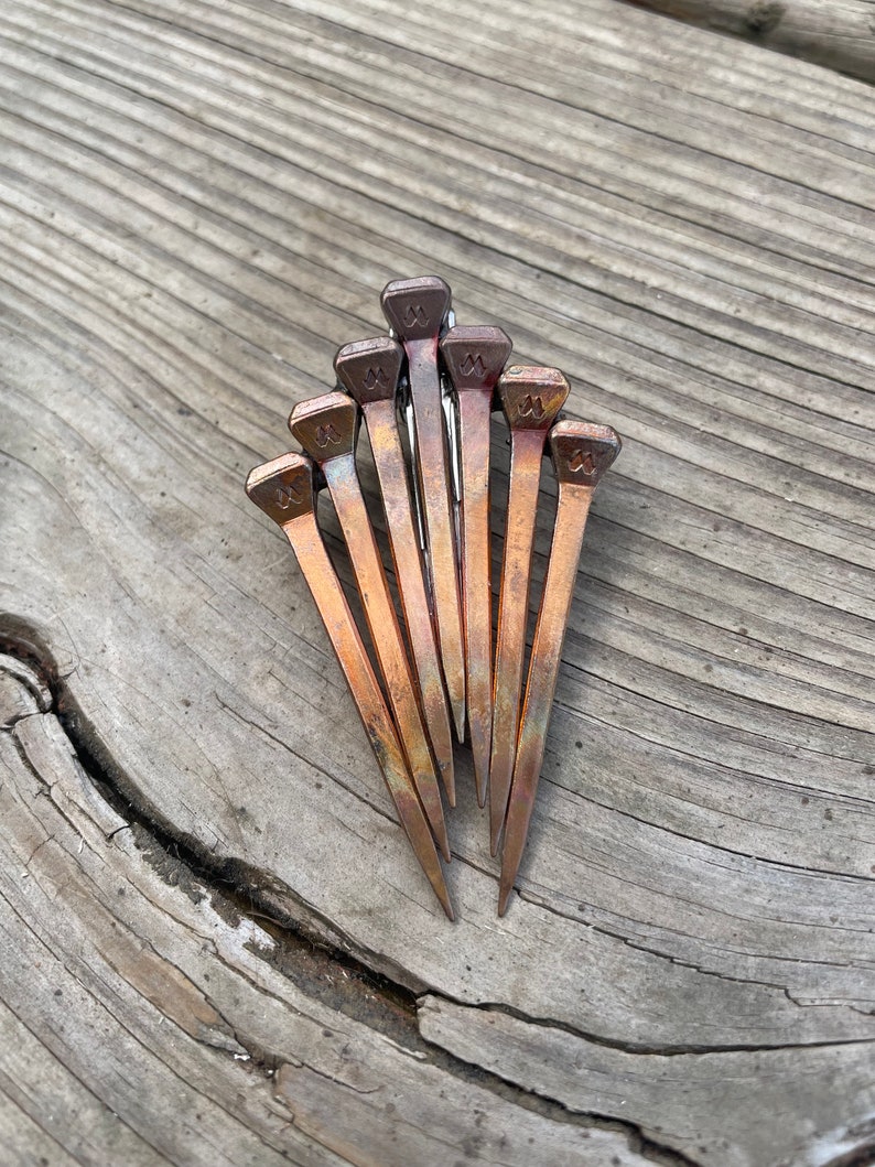 Op de afbeelding: Een verzameling van negen koperkleurige hoefijzernagels, gerangschikt in een waaier. Elke nagel heeft een rechthoekige kop met een decoratief ontwerp. De nagels liggen op een verweerd, grijs houten oppervlak met zichtbare nerven en scheuren.