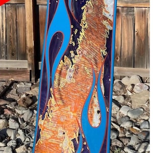 May include: A skateboard deck featuring a vibrant design with bright blue flame patterns outlined in thin orange lines. The centre of the deck displays a textured, mottled pattern in shades of orange, purple, and metallic gold. The text SOLD OUT! is visible.
