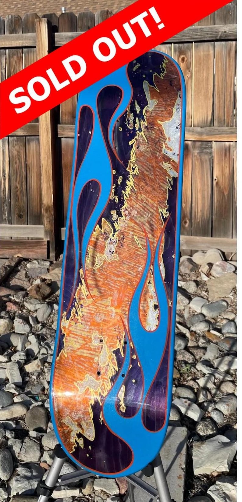 May include: A skateboard deck featuring a vibrant design with bright blue flame patterns outlined in thin orange lines. The centre of the deck displays a textured, mottled pattern in shades of orange, purple, and metallic gold. The text SOLD OUT! is visible.