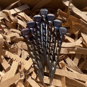 Op de afbeelding: Een cluster van zeven handgemaakte metalen haarspelden. Elke speld heeft een gedraaide schacht en een vierkante kop met een ruitvormig ontwerp. De spelden hebben een donkere, iriserende afwerking en liggen in bruin verpakkingsmateriaal.