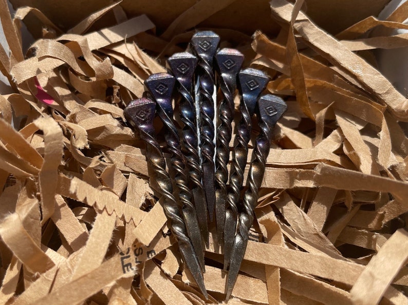 Op de afbeelding: Een cluster van zeven handgemaakte metalen haarspelden. Elke speld heeft een gedraaide schacht en een vierkante kop met een ruitvormig ontwerp. De spelden hebben een donkere, iriserende afwerking en liggen in bruin verpakkingsmateriaal.