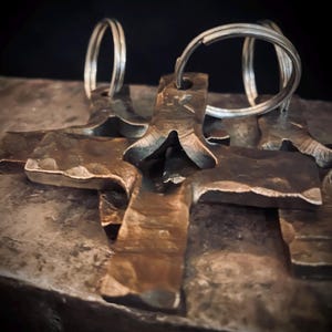May include: Close-up of several metal keychains with a cross design. The keychains are made of a dark, textured metal with a hammered appearance. Each keychain has a silver-colored ring attached for keys.