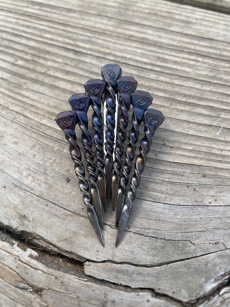 Op de afbeelding: Een handgemaakte metalen sculptuur met zeven donkergrijze spijkers, gerangschikt in een waaier. De spijkers hebben gedraaide schachten en ruitvormige koppen met een decoratief ontwerp. De sculptuur wordt getoond op een verweerd houten oppervlak.