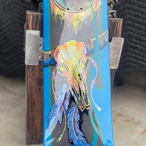 May include: A skateboard deck featuring a central graphic of a multi-coloured animal skull adorned with hanging blue feathers and beads. The background is dark grey with bright blue borders and a circular emblem at the top containing arrows.