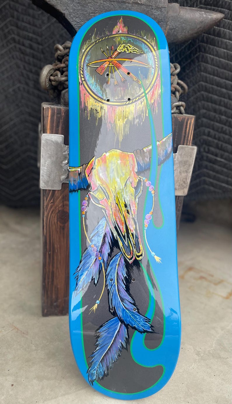 May include: A skateboard deck featuring a central graphic of a multi-coloured animal skull adorned with hanging blue feathers and beads. The background is dark grey with bright blue borders and a circular emblem at the top containing arrows.