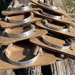 May include: A stack of silver metal bracelets with various inspirational phrases stamped on them, including "breathe", "stay wild", and "choose joy". The bracelets are displayed on brown cardboard packaging with twine.