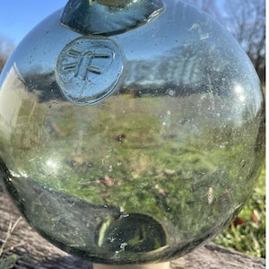 Puede incluir: Una gran boya redonda de vidrio verde con una base blanca. La boya tiene una marca circular con un símbolo.