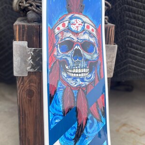 May include: A skateboard deck featuring a graphic illustration of a skull wearing a feathered headdress. The artwork uses a vibrant blue and red colour palette with white accents. The design includes diagonal stripes at the bottom and a white border.