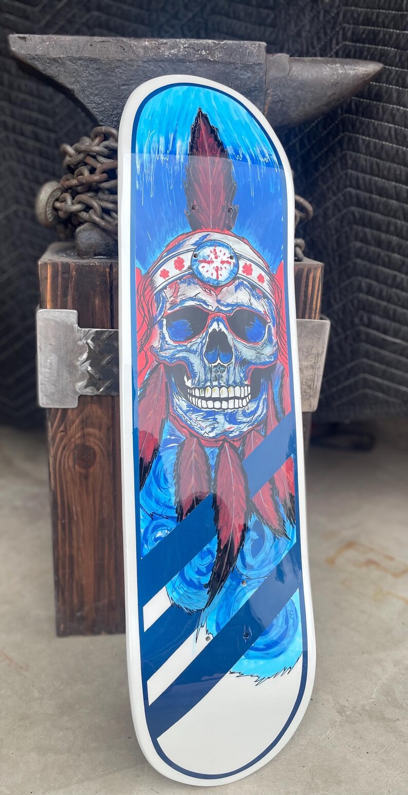 May include: A skateboard deck featuring a graphic illustration of a skull wearing a feathered headdress. The artwork uses a vibrant blue and red colour palette with white accents. The design includes diagonal stripes at the bottom and a white border.