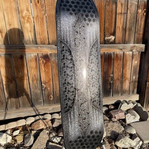 May include: A skateboard deck featuring a black and silver glitter finish with a repeating hexagonal pattern at the top and bottom, and a geometric cube design along the centre sides, all set against a dark, shimmering metallic background.