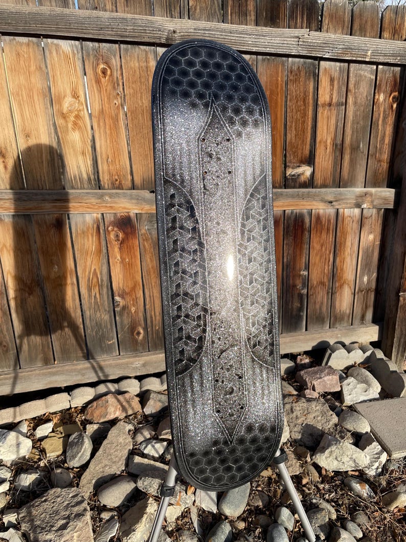 May include: A skateboard deck featuring a black and silver glitter finish with a repeating hexagonal pattern at the top and bottom, and a geometric cube design along the centre sides, all set against a dark, shimmering metallic background.