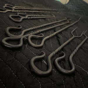 Könnte beinhalten: Ein Set aus dunkelgrauen Metall-Bratgabeln mit langen, schlanken Schäften, gedrehten Details und geschlungenen Griffen. Jede Gabel endet in einer zweizinkigen Spitze und weist ein handgeschmiedetes Aussehen mit einer matten Oberfläche auf.