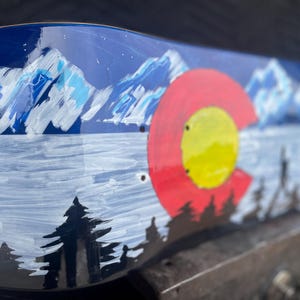 May include: A skateboard deck featuring a hand-painted landscape of snow-capped blue mountains, a dark forest silhouette, and a large red and yellow circular emblem inspired by the Colorado state flag against a deep blue night sky background.