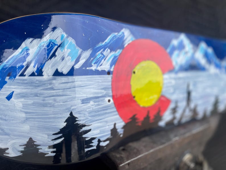 May include: A skateboard deck featuring a hand-painted landscape of snow-capped blue mountains, a dark forest silhouette, and a large red and yellow circular emblem inspired by the Colorado state flag against a deep blue night sky background.