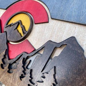 May include: Wooden wall art featuring a layered design of mountains, a setting sun, and the Colorado state flag. The artwork uses shades of brown, red, yellow, and blue, creating a dimensional effect. The piece is likely for home decor.