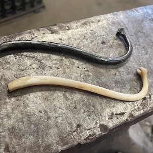May include: Two hooks, one made of metal and one made of bone, are shown on a metal surface. The metal hook is dark grey and the bone hook is light beige.