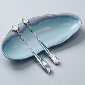 May include: Silver drop earrings featuring a clear, round gemstone and a teardrop pendant with a mother-of-pearl inlay. The earrings are suspended from a delicate chain and presented on a light blue, oval dish.