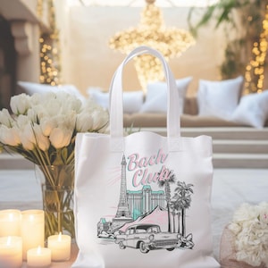 May include: White tote bag with a graphic design featuring the Eiffel Tower, palm trees, a classic car, and the text "Bach Club" in pink and teal. The bag has two white handles.