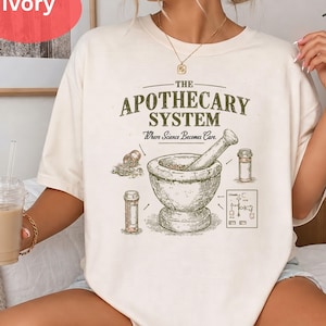 May include: An ivory short-sleeve t-shirt featuring a dark green graphic print. The text reads THE APOTHECARY SYSTEM Where Science Becomes Care. The illustration depicts a mortar and pestle, two medicine bottles, and a small technical diagram.