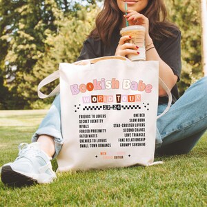 May include: A white tote bag with the text "Bookish Babe World Tour 23-24" in pink, orange, and brown lettering. The bag lists various bookish tropes, such as "Friends to Lovers", "Slow Burn", and "Star-Crossed Lovers".