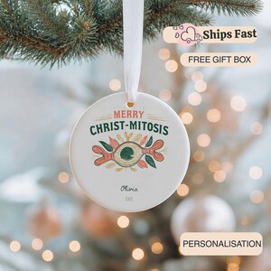 May include: A round white ceramic ornament hanging from a white ribbon. The front features the text MERRY CHRIST-MITOSIS in red and green, with a central illustration of a dividing cell in orange, green, and brown. The name Olivia and DATE are printed below.