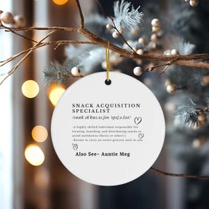 May include: A white, round ceramic ornament with the text "SNACK ACQUISITION SPECIALIST" in black. The text includes a phonetic pronunciation and humorous job description. A gold string is attached for hanging. The ornament is displayed on a branch with decorative elements.