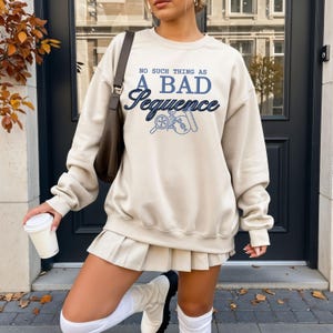 May include: A beige sweatshirt with the text "NO SUCH THING AS A BAD Sequence" in blue. The sweatshirt features a graphic of a magnifying glass, DNA, and a test tube. The person is wearing a pleated skirt and white boots.