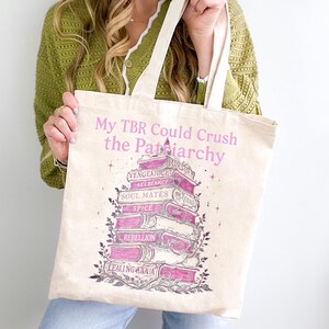 May include: A cream tote bag with the pink text "My TBR Could Crush the Patriarchy". The design features a stack of books with titles such as "Vengeance" and "Rebellion". The bag has cream-colored handles and is made of a light-colored fabric.